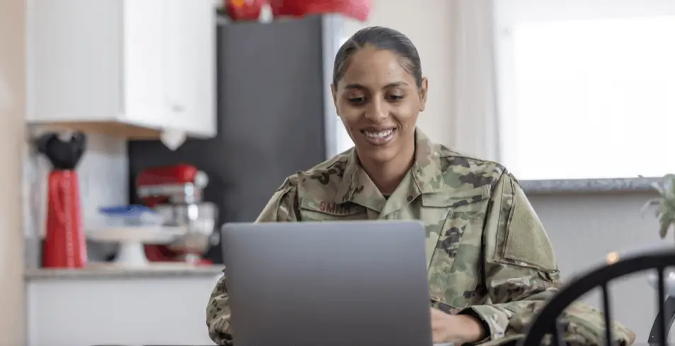 female service member in military fatigues, sitting behind a laptop and smiling