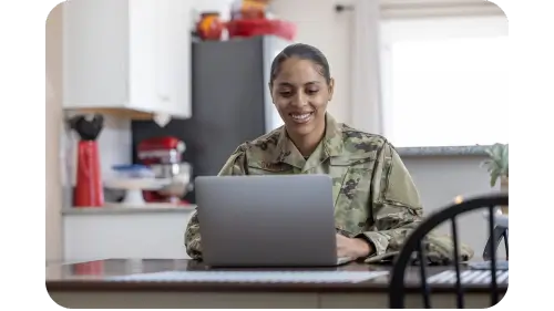 female service member in military fatigues sitting at a table behind a laptop and smiling