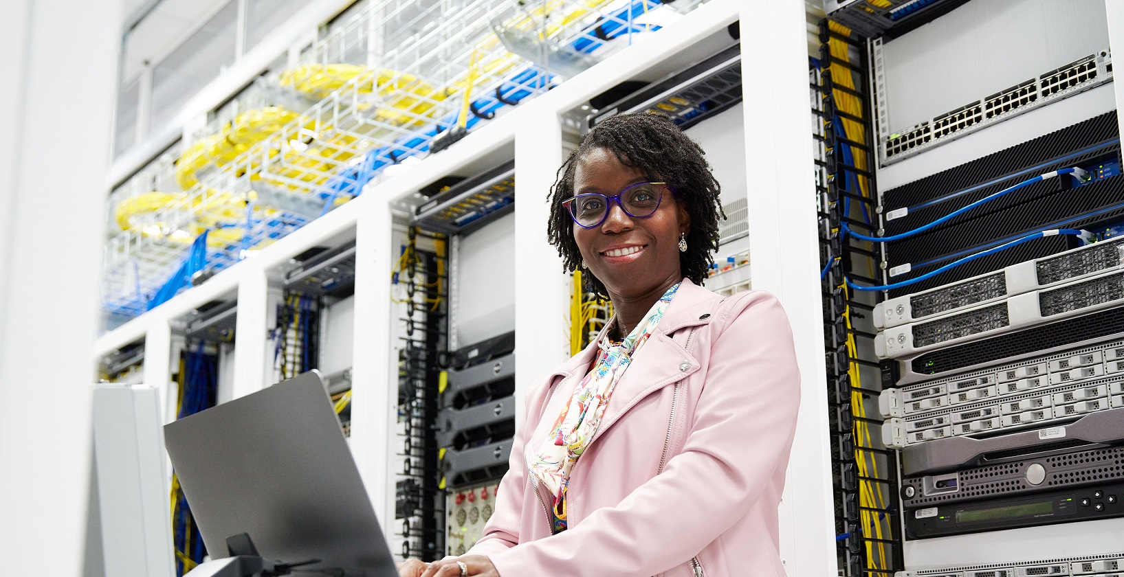 smiling black woman in business attire, standing in front of a computer in a large server room