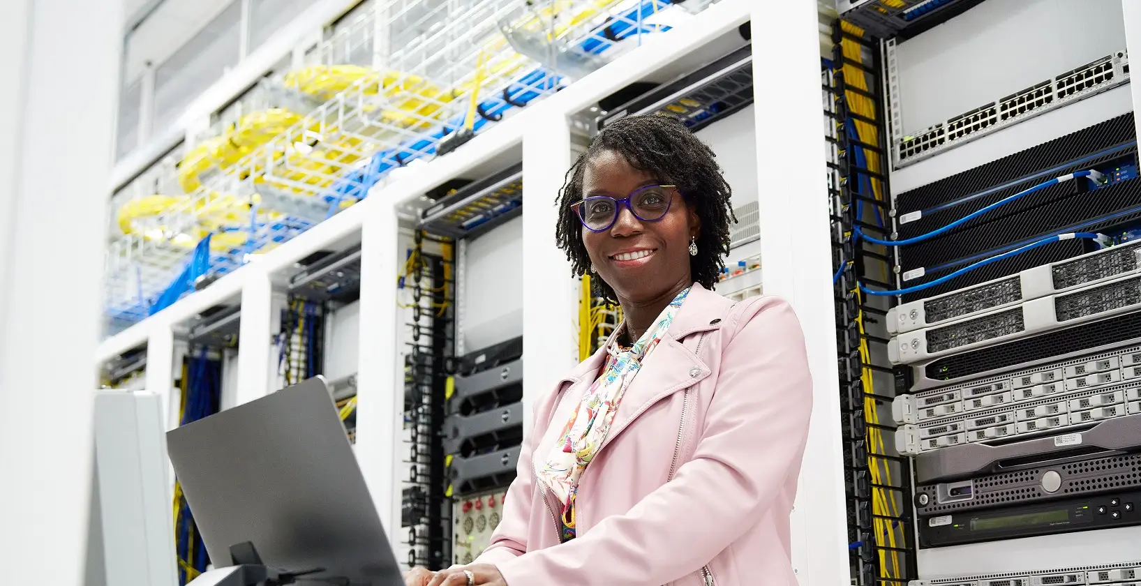 smiling woman in business attire, standing in front of a computer in a large server room
