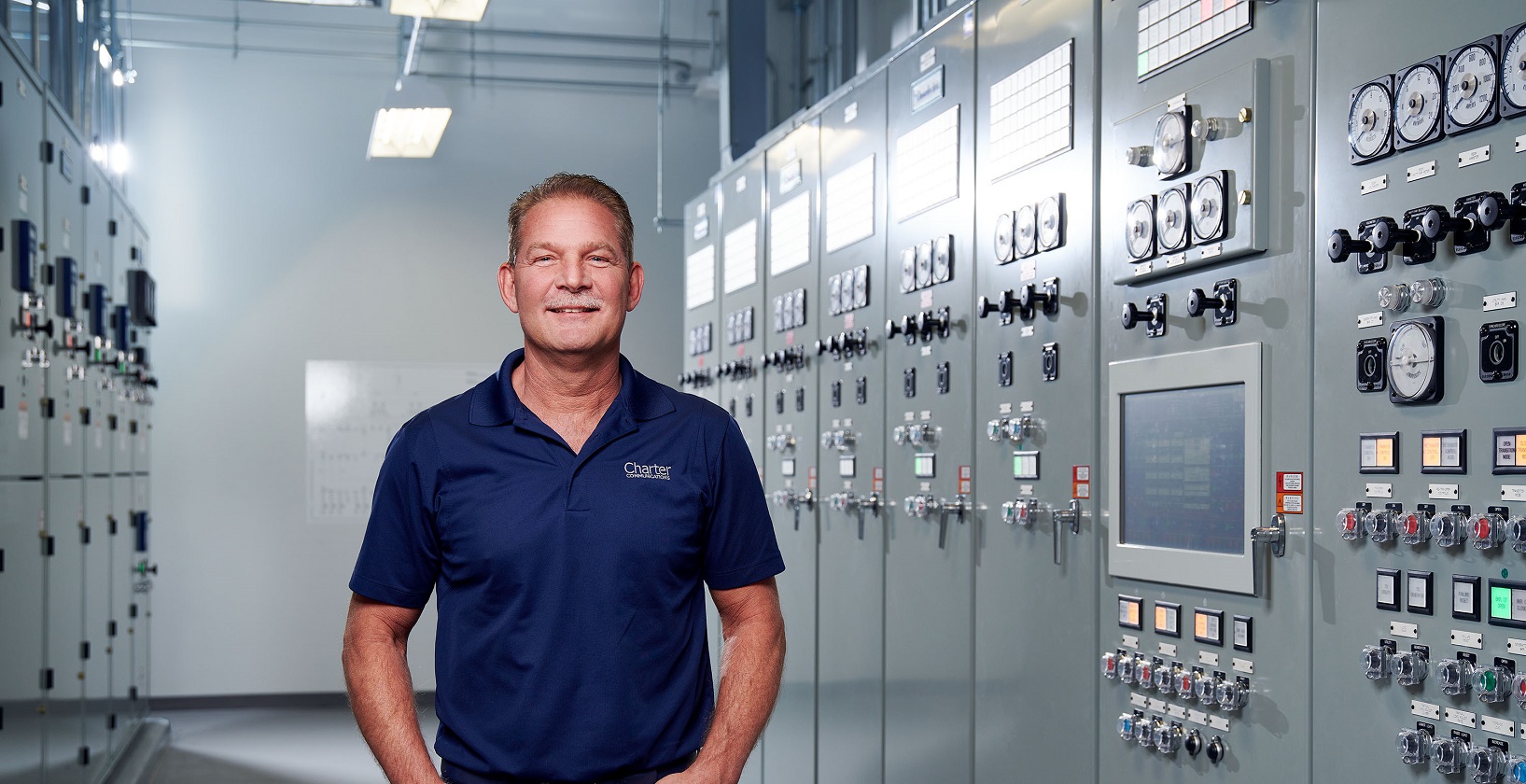 smiling male employee in a dark blue polo shirt, standing in front of a large control panel