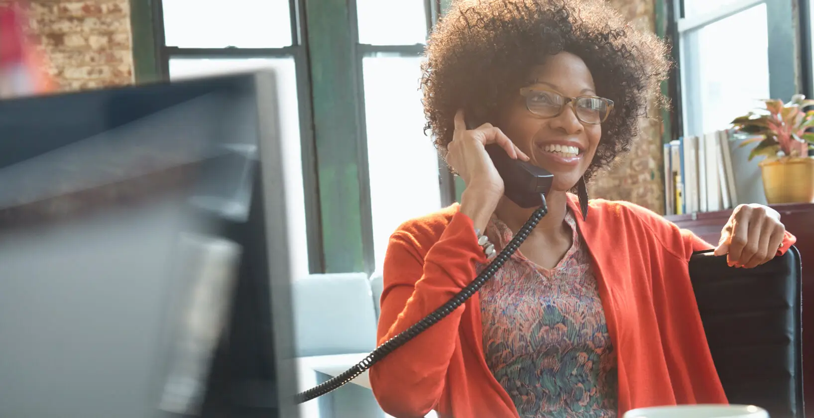 woman in business attire sitting comfortably in a sunlit office, talking on the phone