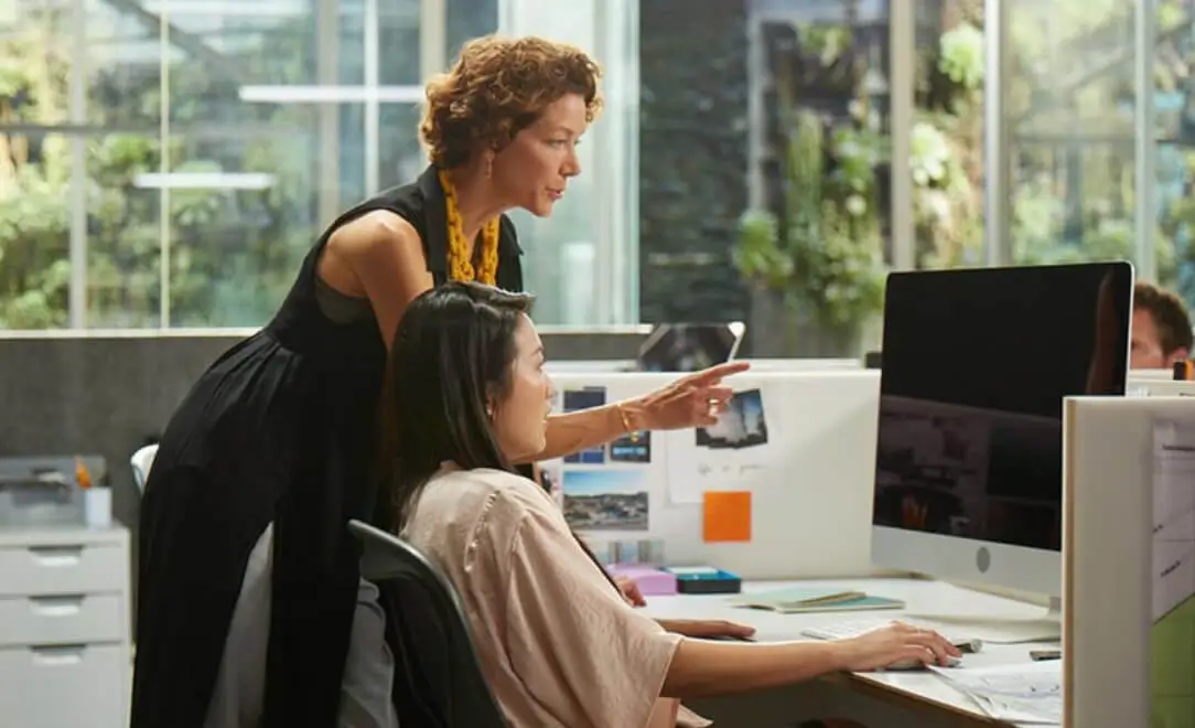 woman in business attire standing over the shoulder of a younger woman sitting at a desk, pointing at the latter's computer screen