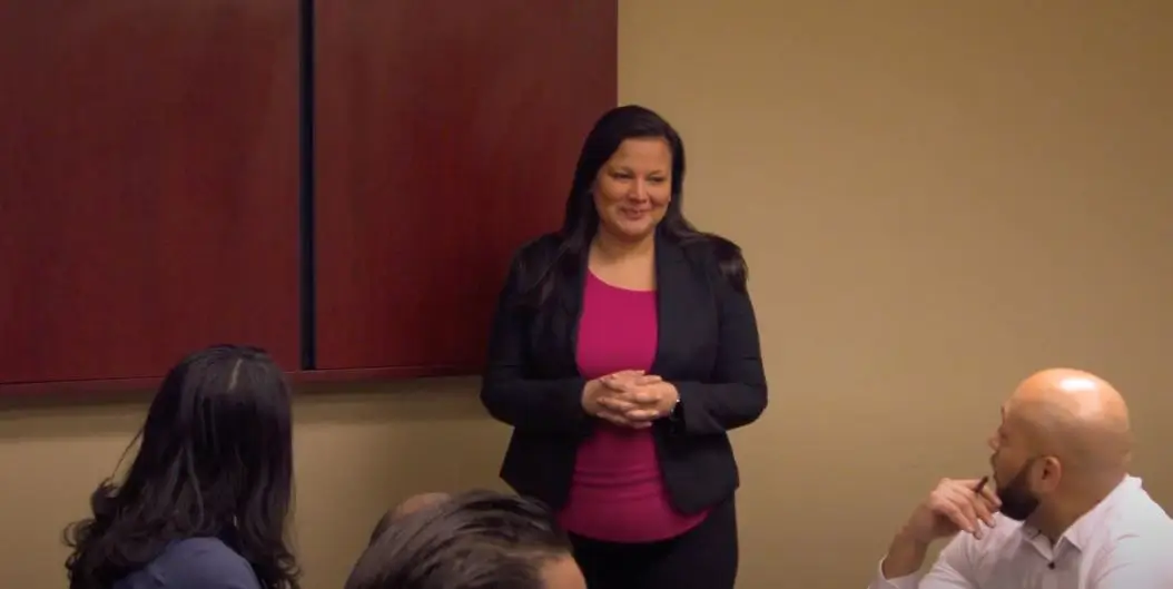 woman in business attire standing in a conference room, speaking to people seated around a table
