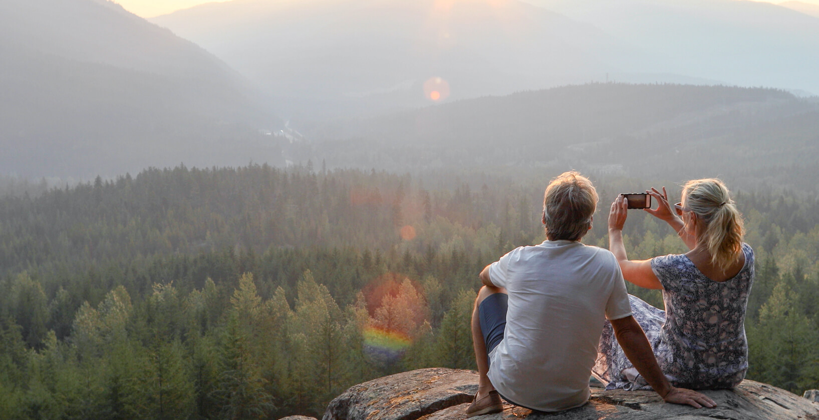 couple sitting on top of a large boulder, taking a photo of the mountains in the distance