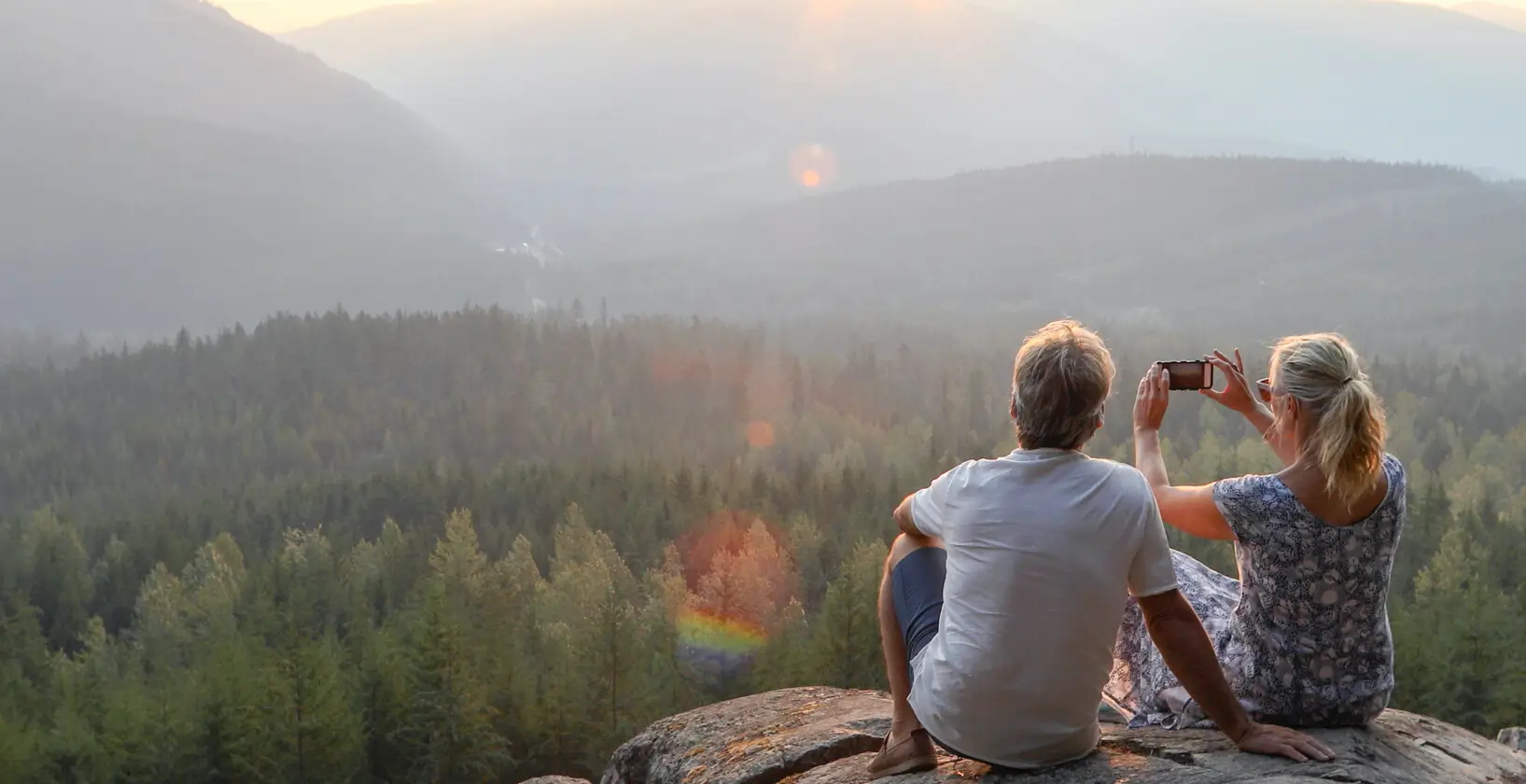 couple sitting on top of a large boulder, taking a photo of the mountains in the distance