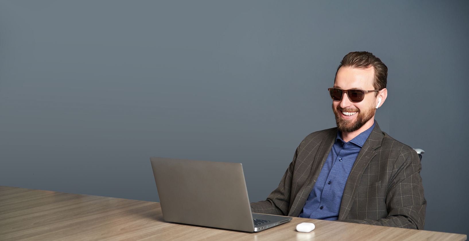 man wearing sunglasses and business attire, seated at a table indoors with a laptop in front of him