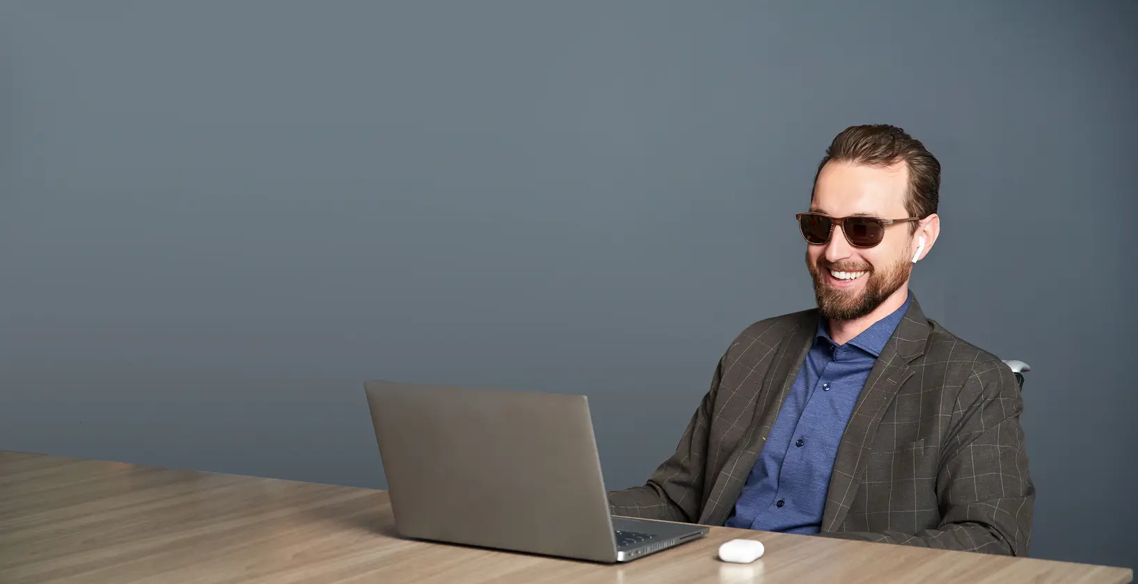 man wearing sunglasses and business attire, seated at a table indoors with a laptop in front of him