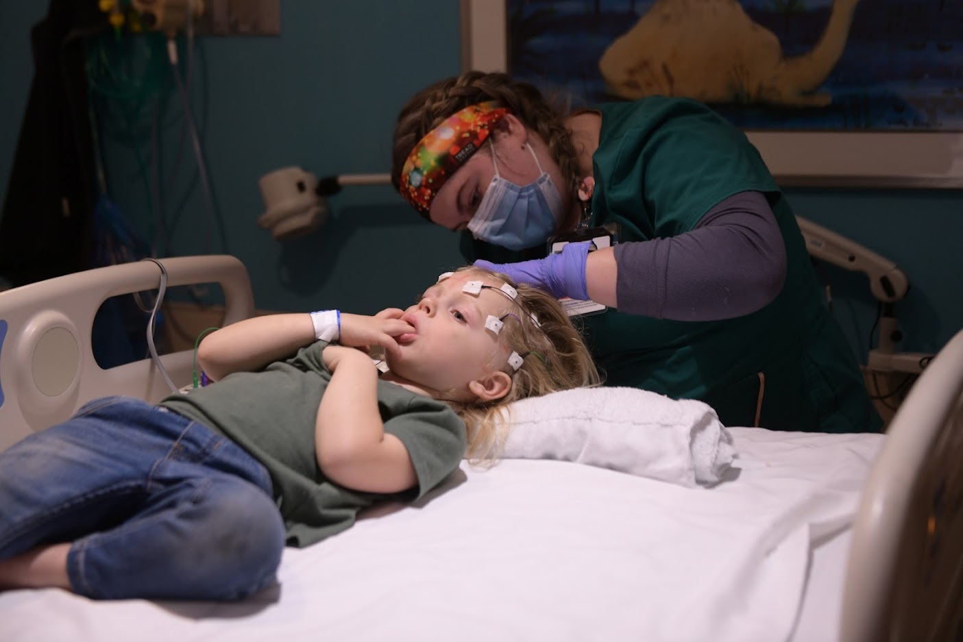 Healthcare professional places monitoring electrodes on a child in a hospital bed during a neurophysiology procedure.