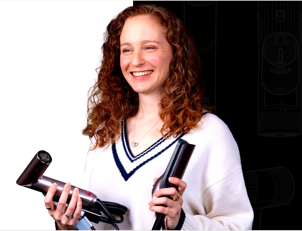 A smiling woman with long, curly auburn hair, wearing a white sweater, holds a metallic hair dryer in one hand and a flat iron straightener in the other.
