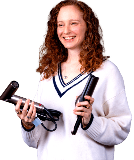 A smiling woman with long, curly auburn hair, wearing a white sweater, holds a metallic hair dryer in one hand and a flat iron straightener in the other.