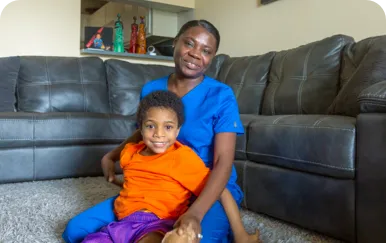 An adult and a child sit on a carpeted floor in front of a dark-colored couch