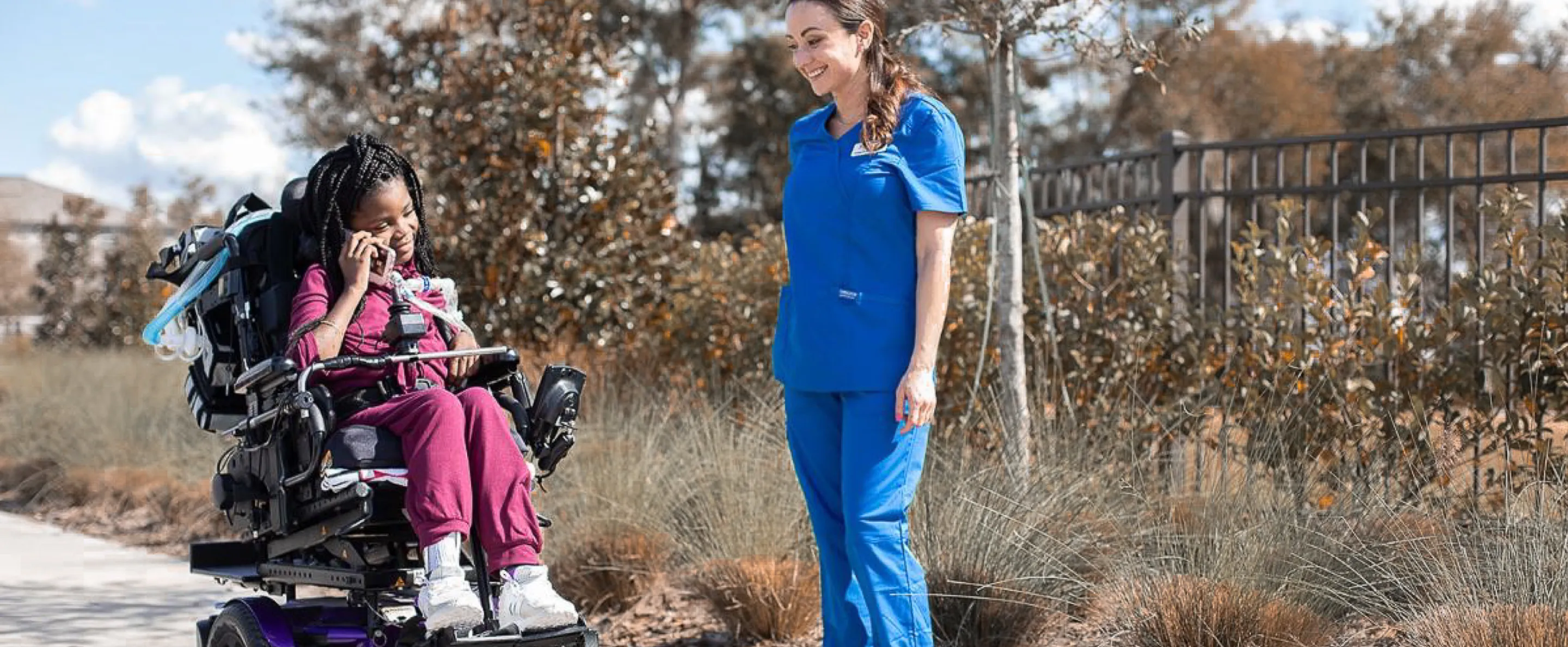 A nurse wearing blue scrubs stands outdoors next to a young girl in a motorized wheelchair.