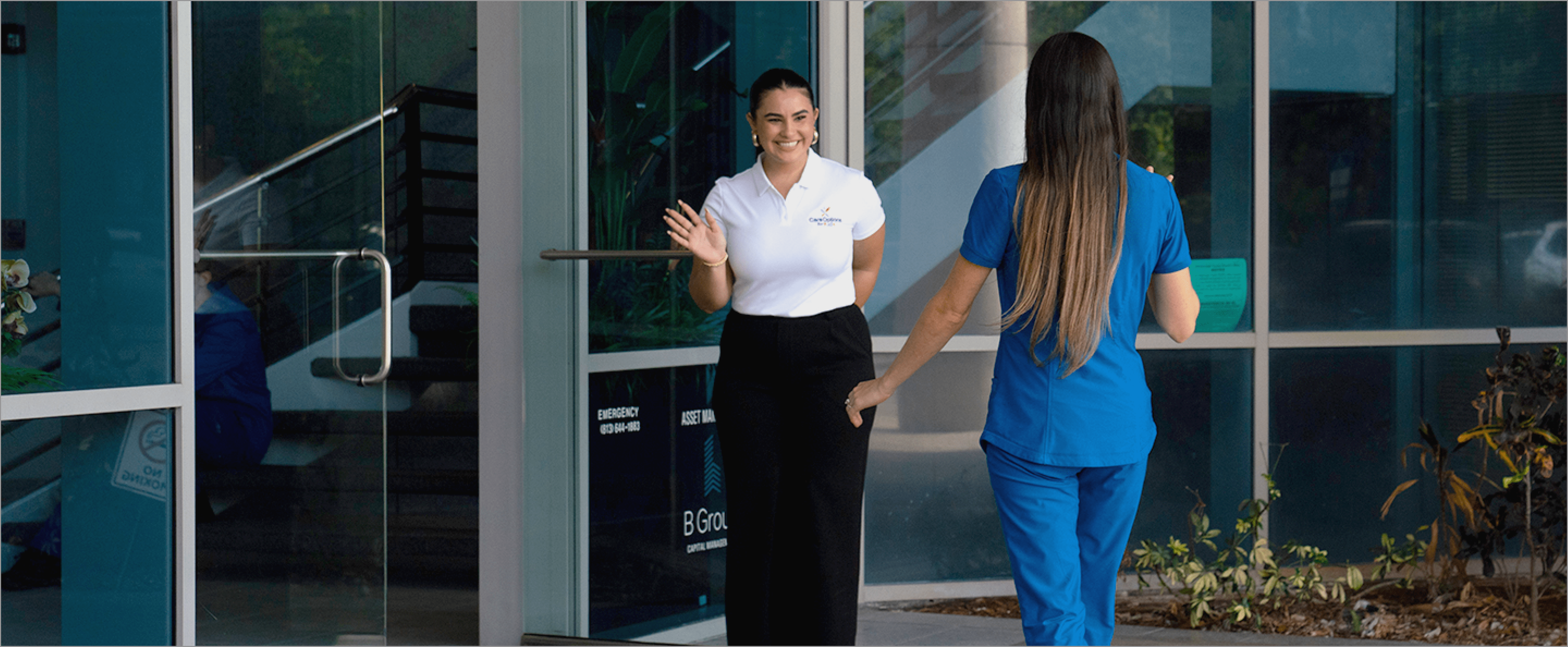 Two women standing outside a modern glass building entrance, one in a white shirt greeting another in blue scrubs, representing a welcoming healthcare facility.