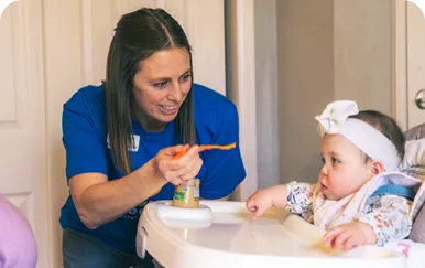 An adult feeds a baby seated in a high chair, guiding a spoon toward the baby.