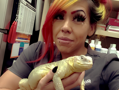Veterinary team member with colorful red, orange, and yellow hair holds a light-colored lizard in a clinic setting. The team member smiles while gently supp
