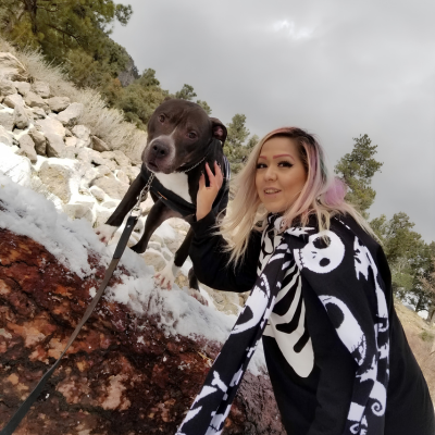 Veterinary hospital team member poses outdoors with a dog during a snowy hike, standing beside a large rock surrounded by snow-covered terrain and trees.
