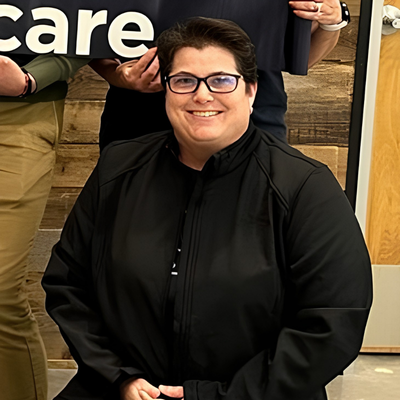 Smiling Petco team member wearing glasses and a black jacket, seated indoors.