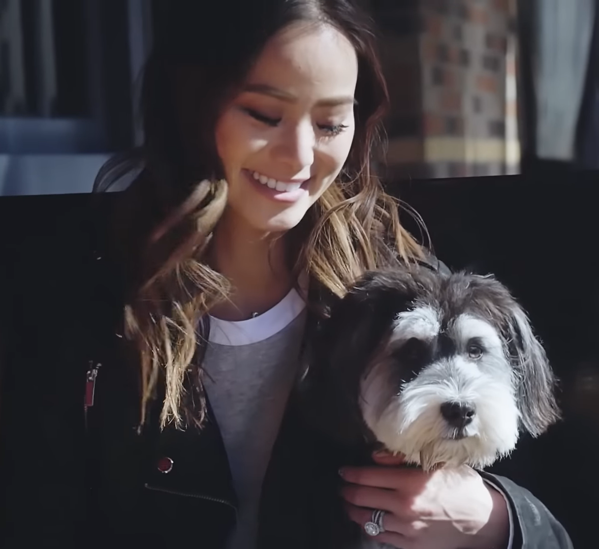 Person with long hair holding a small black and white fluffy dog, wearing a dark jacket and light shirt, with brick and window architecture in the background.