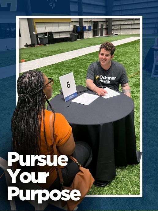 Two people sit across from each other at a round table inside an indoor sports facility, reviewing paperwork next to a sign marked "6." Text reads, "Pursue Your Purpose."