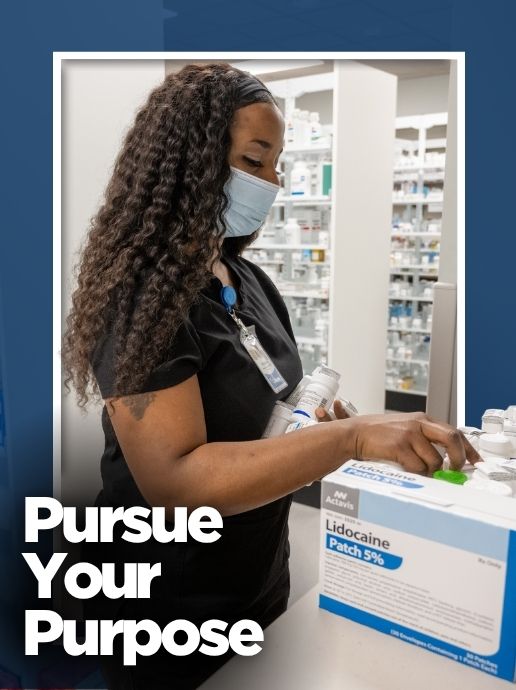 A woman wearing black medical scrubs and a face mask selects medication boxes at a pharmacy workstation. Text reads, "Pursue Your Purpose."