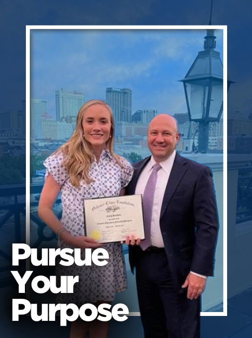 A young woman in a patterned white dress holds a scholarship certificate while standing next to an older man in a suit with a city skyline in the background.