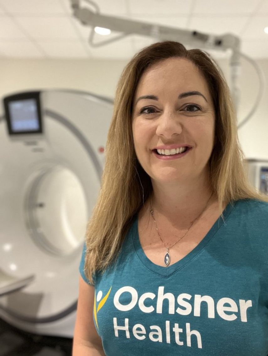 A smiling women in a teal colored Ochsner Health T shirt in front of a CT machine.