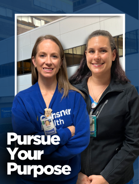 Two smiling women, one in a blue Ochsner Health sweatshirt and the other in a black jacket, stand side-by-side in front of a modern building.