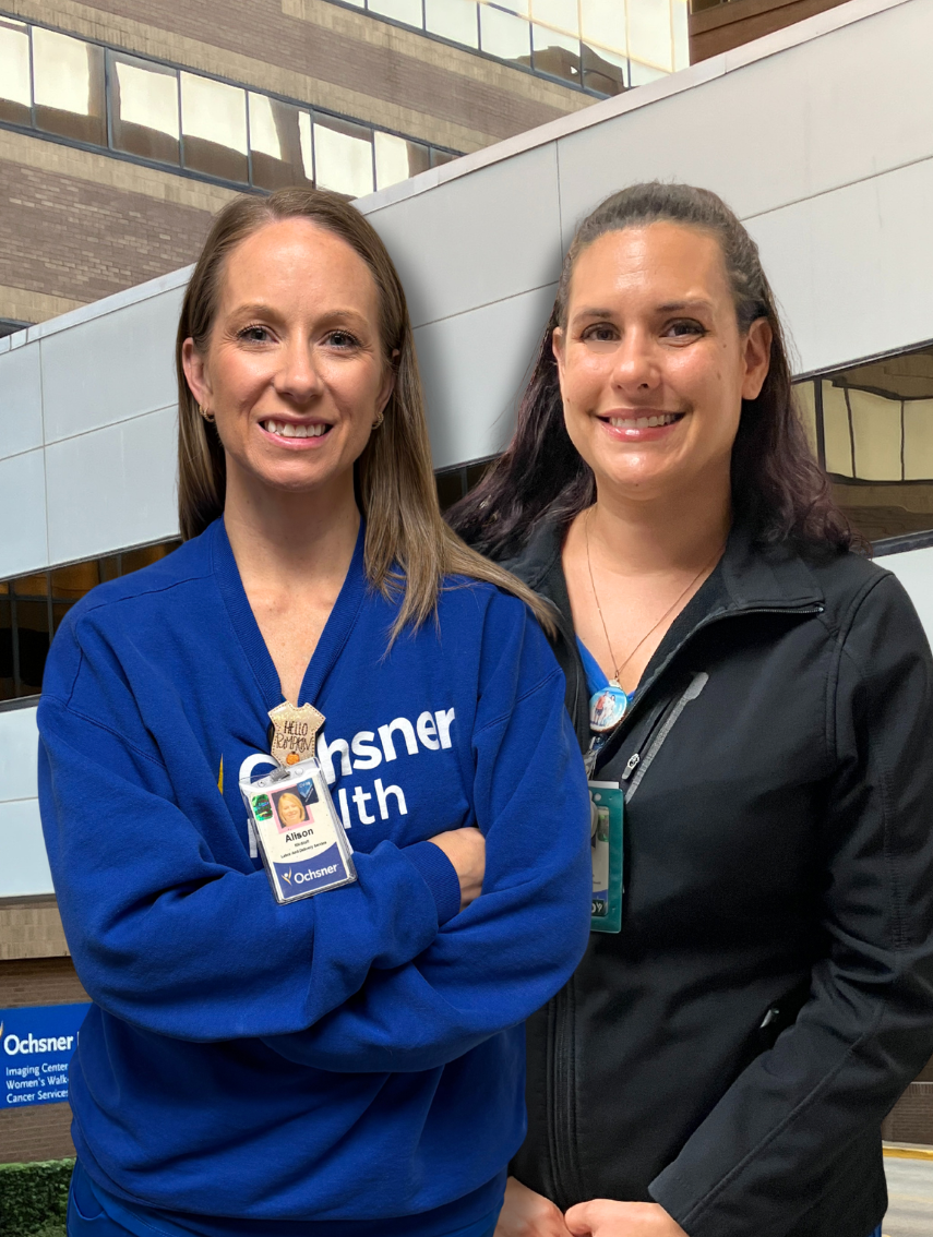 Two smiling women, one in a blue Ochsner Health sweatshirt and the other in a black jacket, stand side-by-side in front of a modern building.