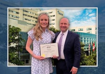 A man and a woman stand in front of a hospital building, with the woman holding a framed certificate.