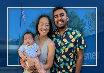 A man and a woman holding an infant stand together in front of a medical building.