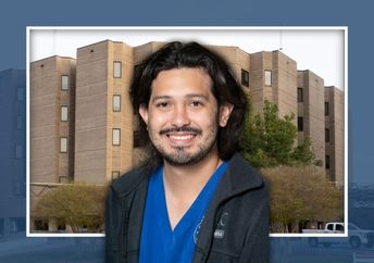 A man wearing blue medical scrubs and a black jacket stands in front of a hospital building.