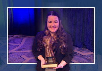 Smiling woman with long dark hair holds a bronze trophy, standing against a backdrop of dark curtains and blue-green stage lighting.