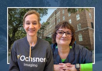 Two women, one in a gray sweatshirt and one in a blue and green layered top, in front of a hospital