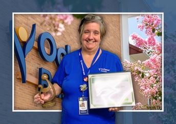 A woman wearing blue medical scrubs holds a framed certificate and a trophy in front of an Ochsner sign.