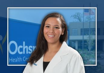 A woman wearing a white coat stands in front of a hospital building and an Ochsner Health Center sign.