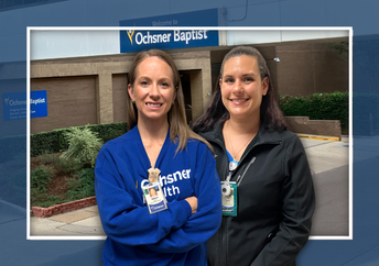 Two smiling women, one in a blue Ochsner Health sweatshirt and the other in a black jacket, stand side-by-side in front of a modern building.