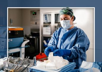 A person wearing a surgical mask, gown, and gloves prepares instruments at a workstation.