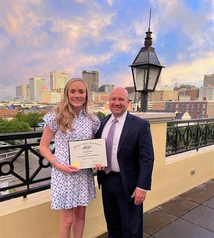 A young woman in a patterned white dress holds a scholarship certificate while standing next to an older man in a suit with a city skyline in the background.