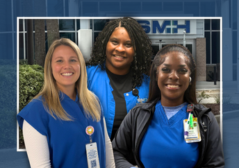 Three smiling female healthcare professionals in blue scrubs stand in front of a modern hospital building with large glass windows and a palm tree.
