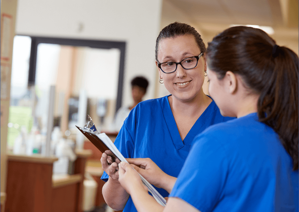 Two nurses in blue scrubs looking at a clipboard