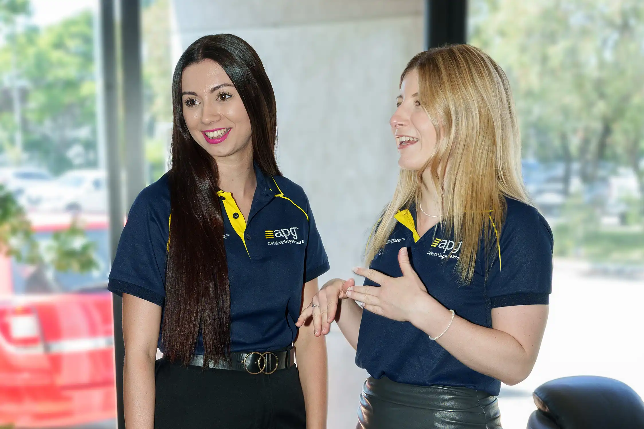 Two women wearing APG uniforms smile and talk while standing indoors, representing career growth and professional development.
