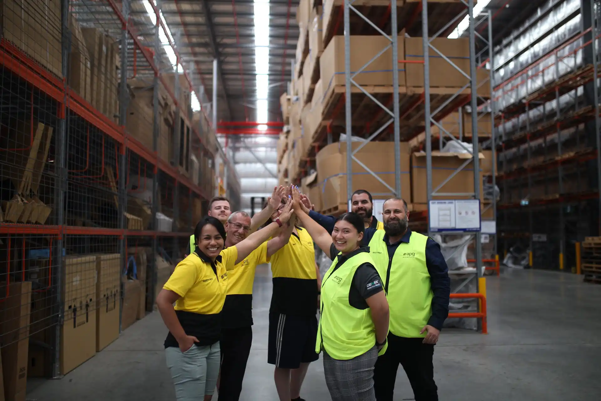 A team of APG employees standing together in a warehouse, smiling and waving, highlighting diversity and team collaboration