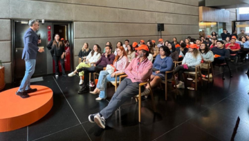 A presenter on stage in front of a group of dozens of SCJ people sitting to watch the presentation