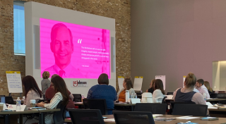 A room of participants looking toward the front of the room where someone is presenting with a slide featuring SCJ's This We Believe values quote