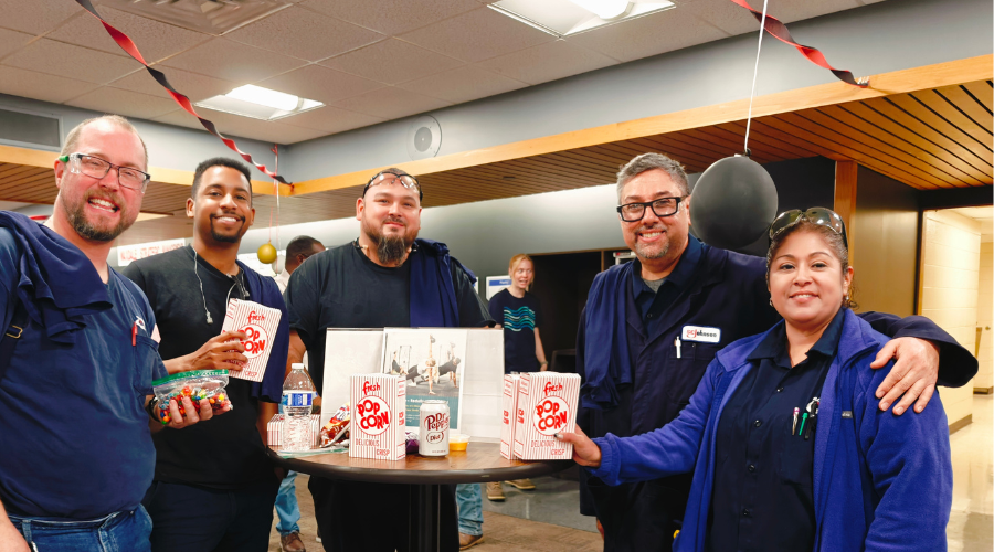 Five team members pose for a group photo around a table with snacks and balloons at appreciation event