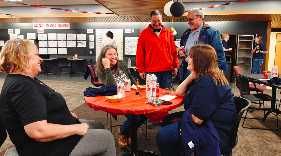 Five team members pose for a group photo around a table with snacks and balloons at appreciation event