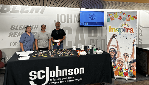 A display table for SC Johnson has several people smiling behind it, and is covered with featured items and next to an Inspira sign.