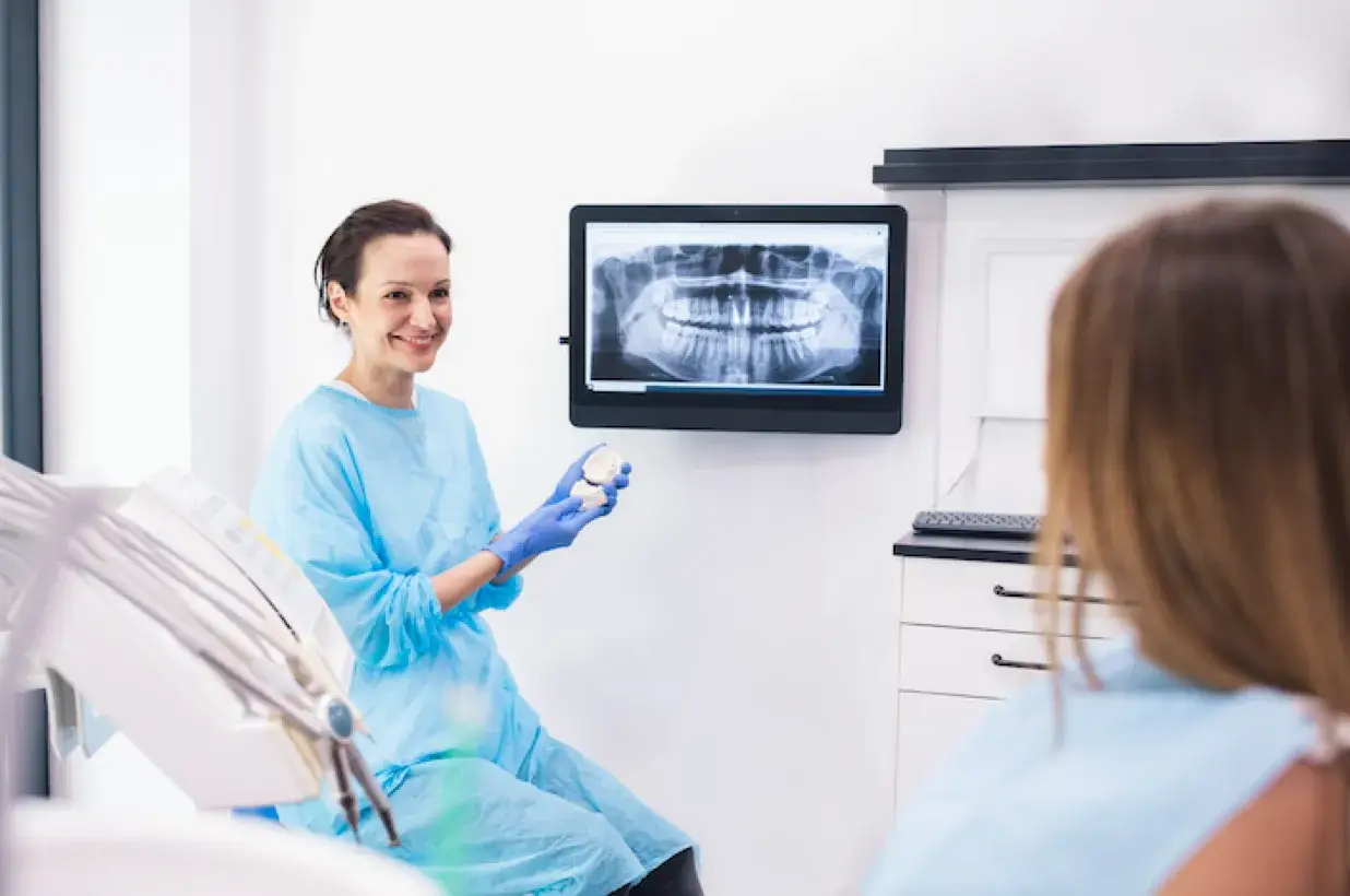 female dental assistant holding a model of a patient's teeth in front of an x-ray image on a screen and smiling at the seated patient