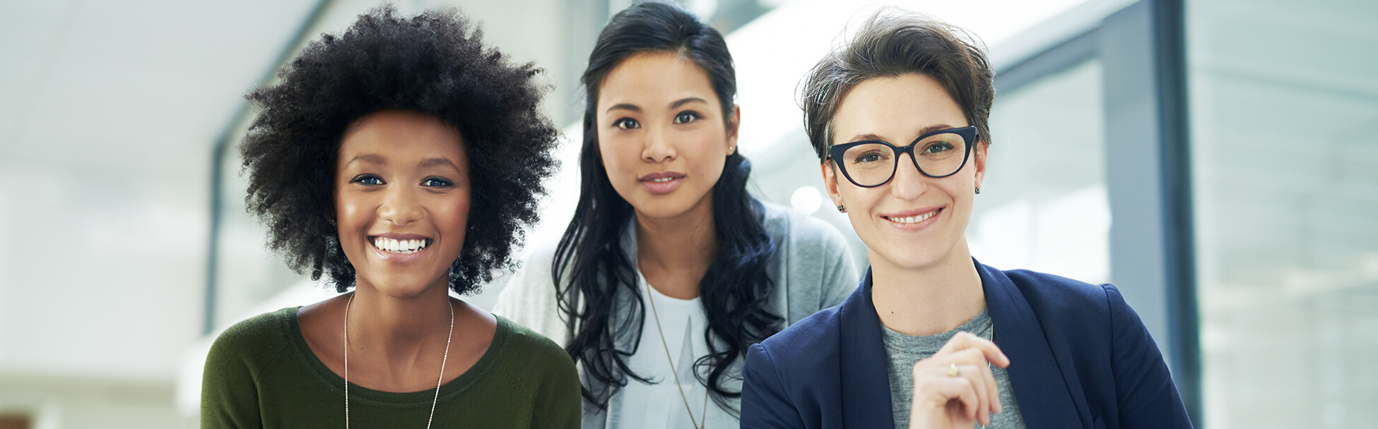 Three smiling racially diverse women in a work environment.